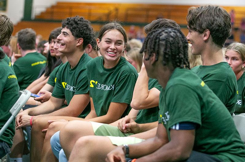 Students wearing matching green shirts that says "Skidmore" great each other while seated in a gymnasium